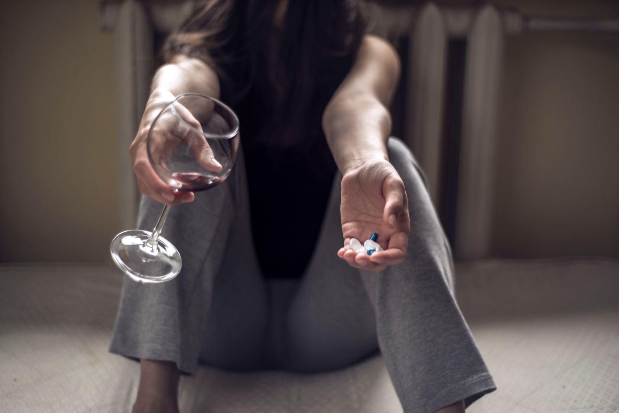 a young girl sitting in a dirty room, with a glass of wine, and holds pills in her hand.