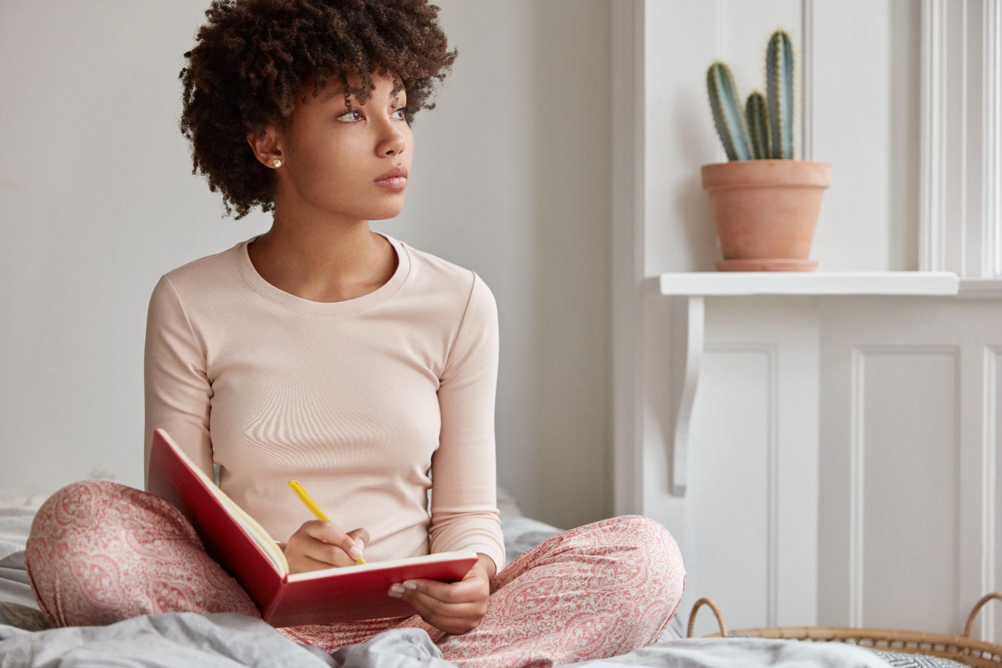 Dark skinned woman sits crossed legs, wears pyjamas, makes notes in diary