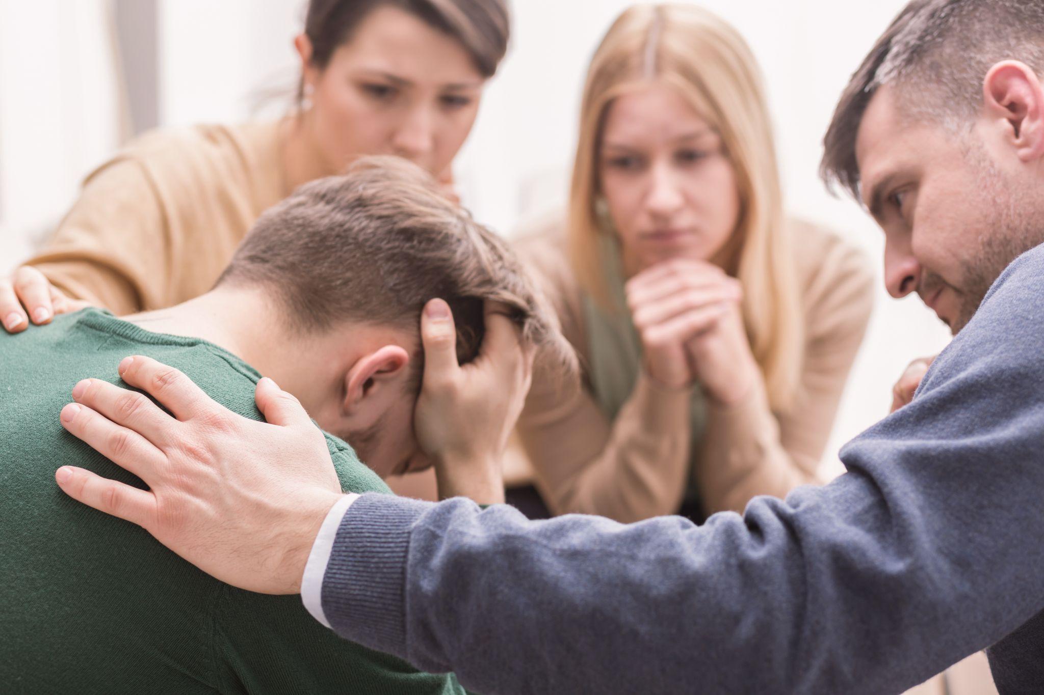 young man holding his head in his hands and friends supporting him during group therapy