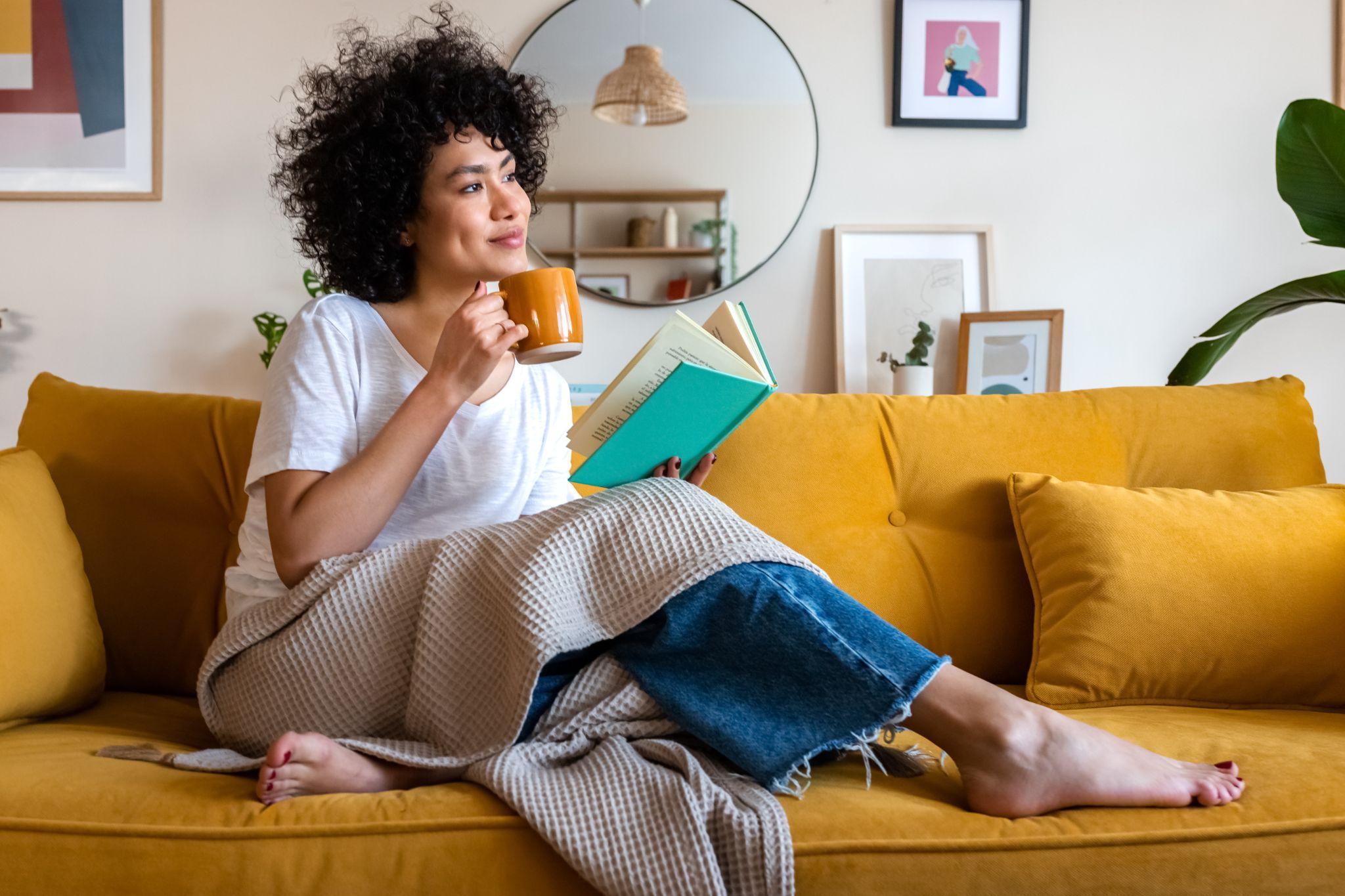 African american woman reading a book at home, drinking coffee sitting on the couch
