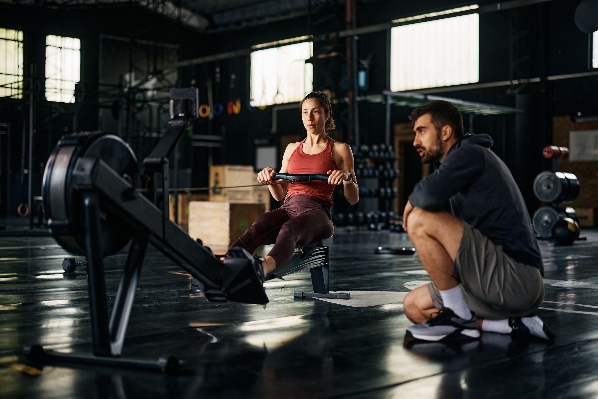 Athletic woman exercising on rowing machine while having cross training with fitness instructor at gym.