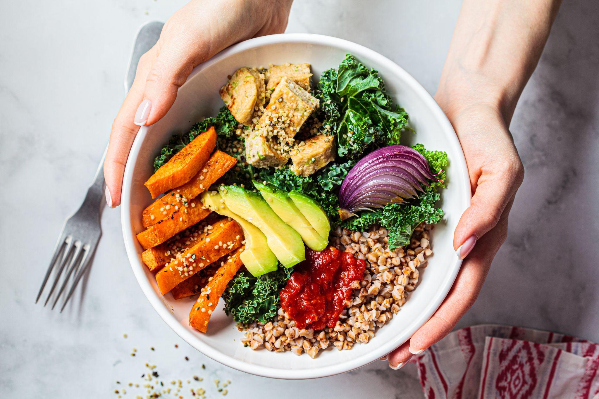 Woman hands eating vegan salad of baked vegetables, avocado, tofu and buckwheat buddha bowl, top view