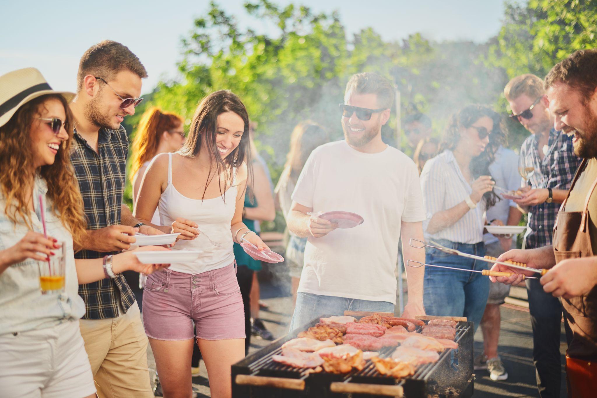 Group of people standing around grill, chatting, drinking and eating
