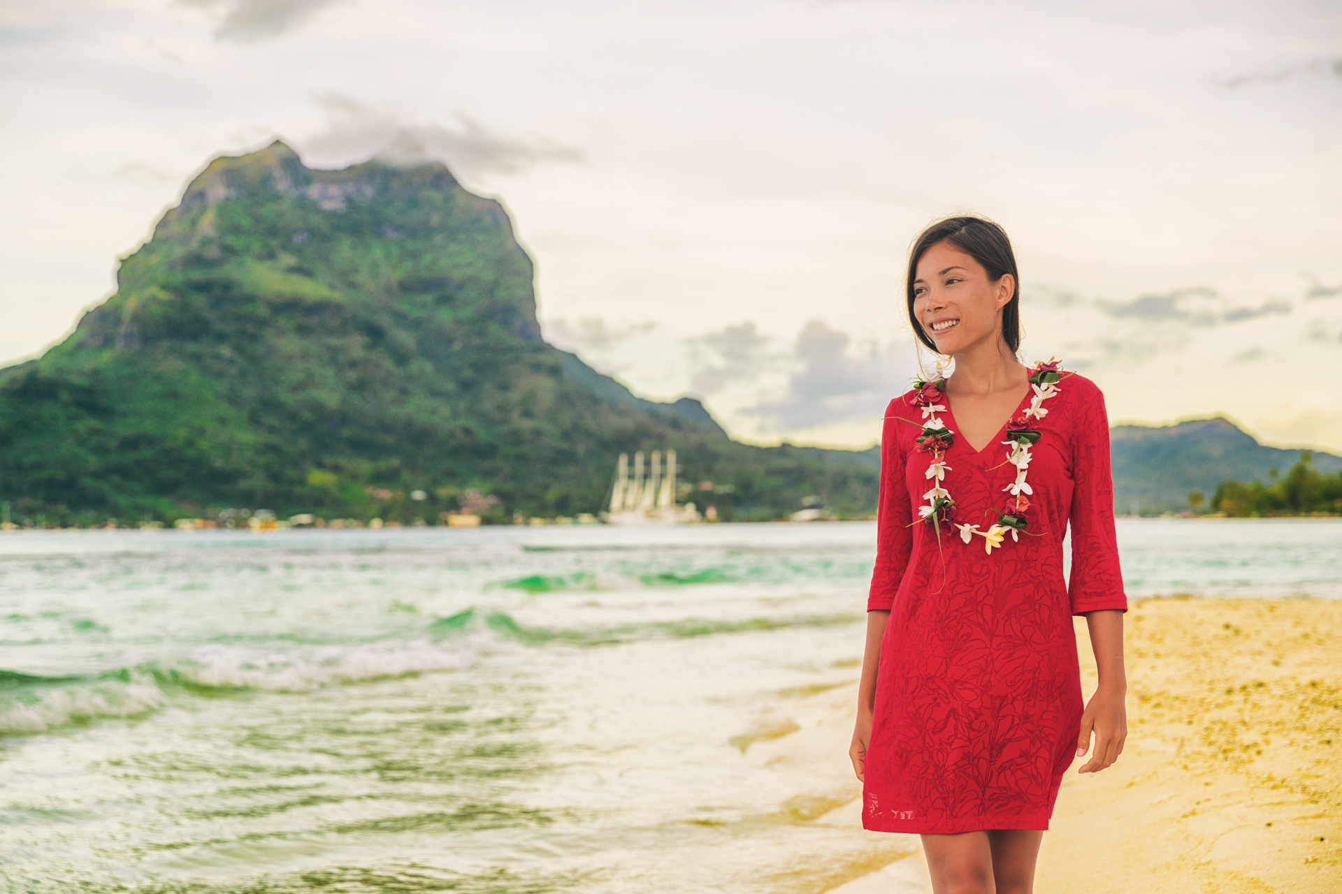 Woman in loose red dress and lei walking down a beach in Hawaii.